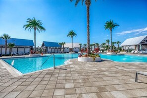 A swimming pool area with multiple pools, palm trees, lounge chairs, and a tiled walkway.