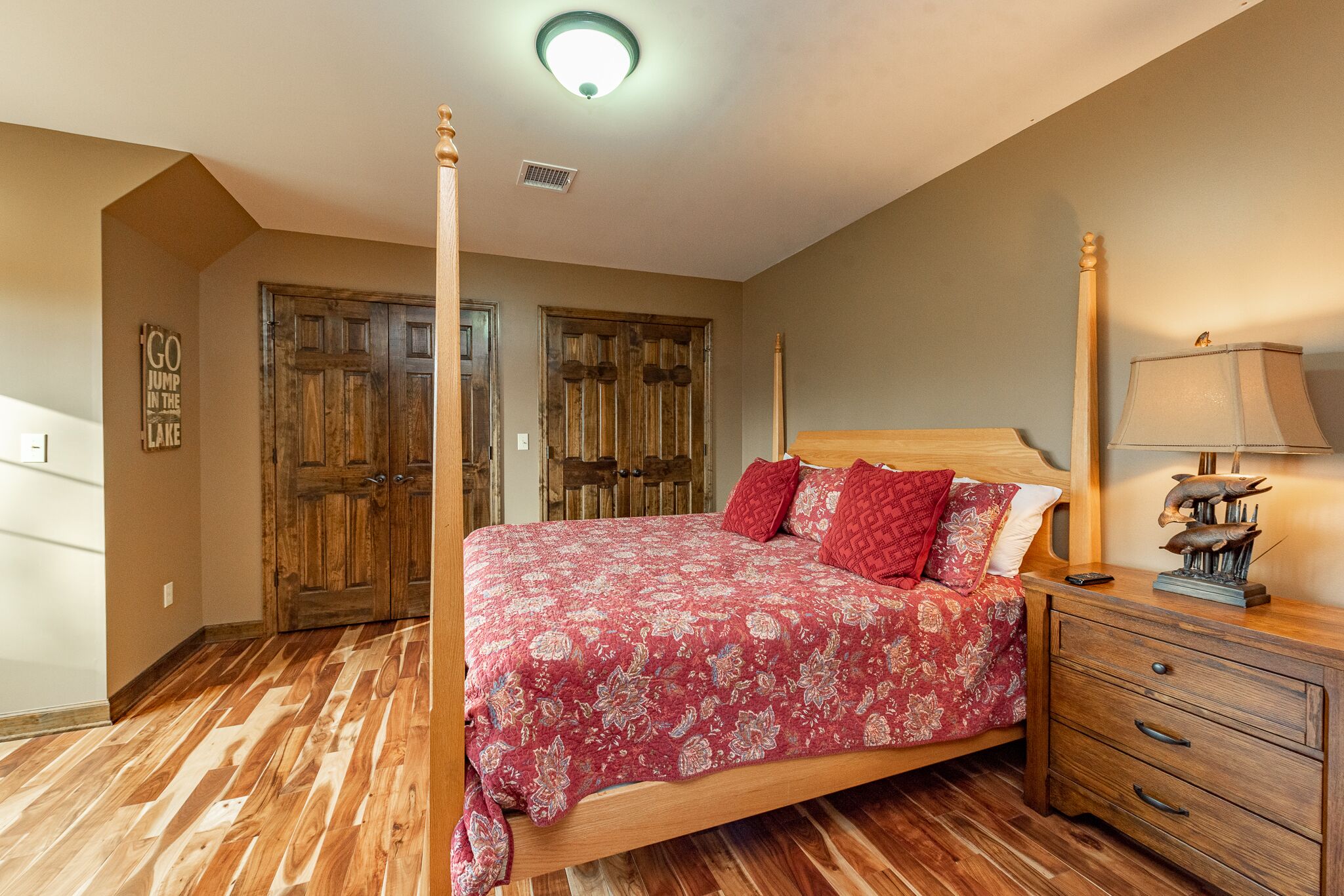 A bedroom with a four-poster bed, wooden flooring, a nightstand with a lamp, and two doors.