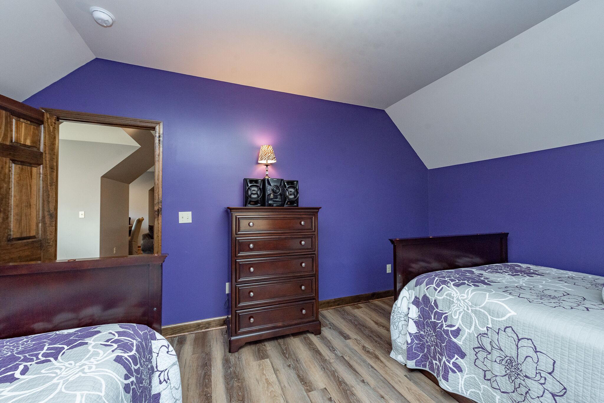 A bedroom with purple walls, a wooden dresser, two beds with floral bedding, and a ceiling light.