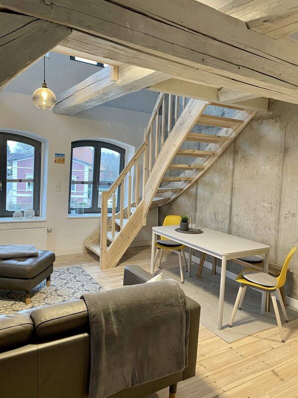 A loft-style room with exposed wooden beams, a staircase, a dining area, and large windows.