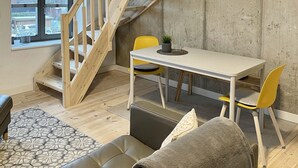 Ein Loft-Zimmer mit freiliegenden Holzbalken, einer Wendeltreppe, einem Essbereich mit Tisch und Stühlen, einem Ledersofa und Blick durch ein großes Fenster.