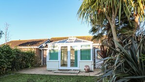 A small, white, single-story house with a glass door and windows, surrounded by a grassy yard and a palm tree. - Dunes View (Newquay)