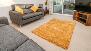 A living room with a grey sofa, yellow cushions, a brown rug, a wooden TV stand, and a sliding glass door to the outside. - Dunes View (Newquay)