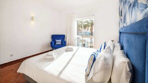 A hotel room with a large bed, a blue armchair, and a view of palm trees through the window. - Villa Loria - beautiful panoramic views (Albufeira)