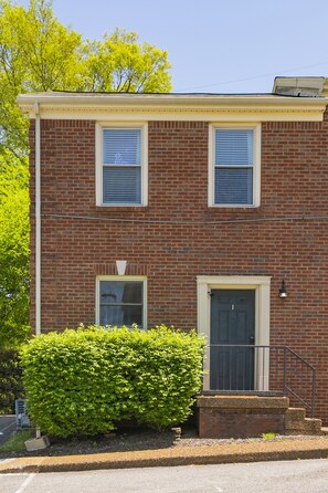 A two-story brick building with a front door, two windows with shutters, and a small front yard with a bush.