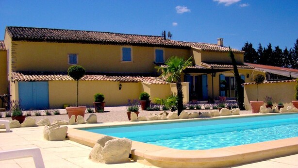 Zona junto a la piscina con piscina, sillas de descanso blancas y un edificio con tejado de azulejos y plantas en macetas.