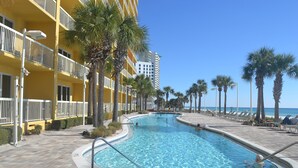 Uma piscina com deck revestido, rodeada por palmeiras e um edifício com varandas.