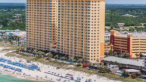 Um hotel alto com várias varandas, uma praia com guarda-sóis e vista para a paisagem ao redor.