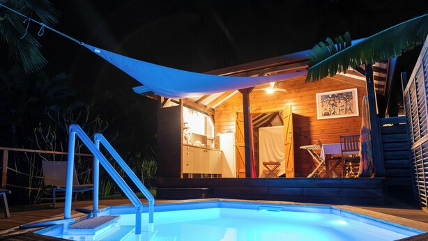 Ein Pool mit Holzterrasse, ein überdachter Sitzbereich im Freien sowie ein Holzgebäude mit Esstisch und Stühlen.