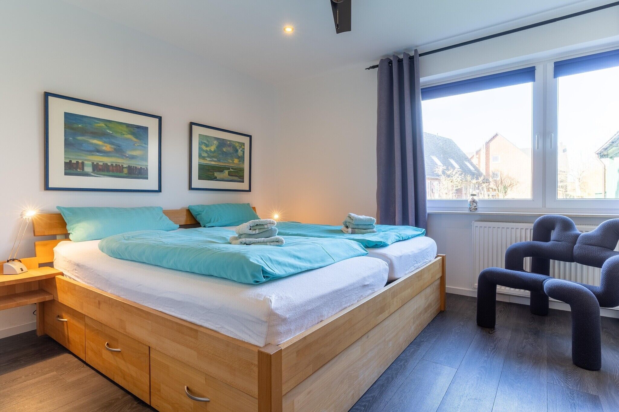 Un dormitorio moderno con una cama de madera, dos cuadros enmarcados de paisajes, una ventana con cortinas y una mesita de noche con lámpara.