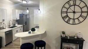 A modern kitchen with a central island, pendant lights, a large wall clock, and a small side table with books. - Riverside Riviera at Seminole Heights (Tampa)