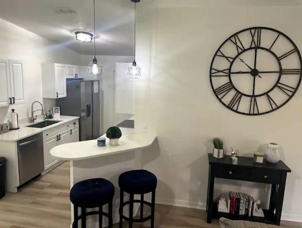 A modern kitchen with a central island, pendant lights, a large wall clock, and a small side table with books.
