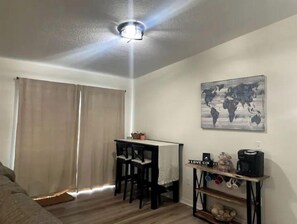 A small, cozy room with a wooden floor, a desk with two chairs, a world map artwork, and a coffee machine.