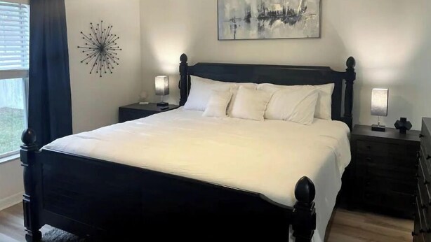 A bedroom with a dark wooden bed, two bedside tables with lamps, a ceiling fan, a window with curtains, and a wall-mounted abstract painting. - Riverside Riviera at Seminole Heights (Tampa)