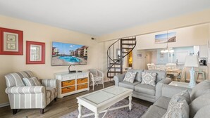 Ein Wohnzimmer mit einer Couch, Sesseln, einem Couchtisch und einem Fernseher. Eine Treppe führt in einen anderen Raum, ein Essbereich mit Tisch und Stühlen sowie eine Küchenzeile mit Arbeitsfläche und Geräten.