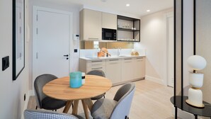 A modern kitchen with white cabinets, a black microwave, and a sink. There is a dining area with a round wooden table and grey chairs. - iloftmalaga Muelle Uno VII (Málaga)
