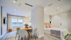 Una habitación de hotel moderna con zona de comedor, una cama con colcha blanca y rosa, y un ventanal con cortinas.
