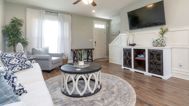 A living room with a ceiling fan, a flat-screen TV mounted on the wall, a round coffee table, a sofa, a dining table, and a cabinet with decorative items.