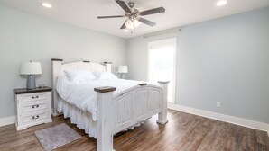 A bedroom with a white bed, wooden floor, ceiling fan, and two bedside tables with lamps.