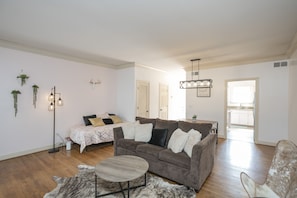 Un salón espacioso con un sofá gris, una mesa de centro y una alfombra. Se observa una cama con edredón estampado y una mesita de noche con lámpara. El suelo es de madera y las paredes son blancas.