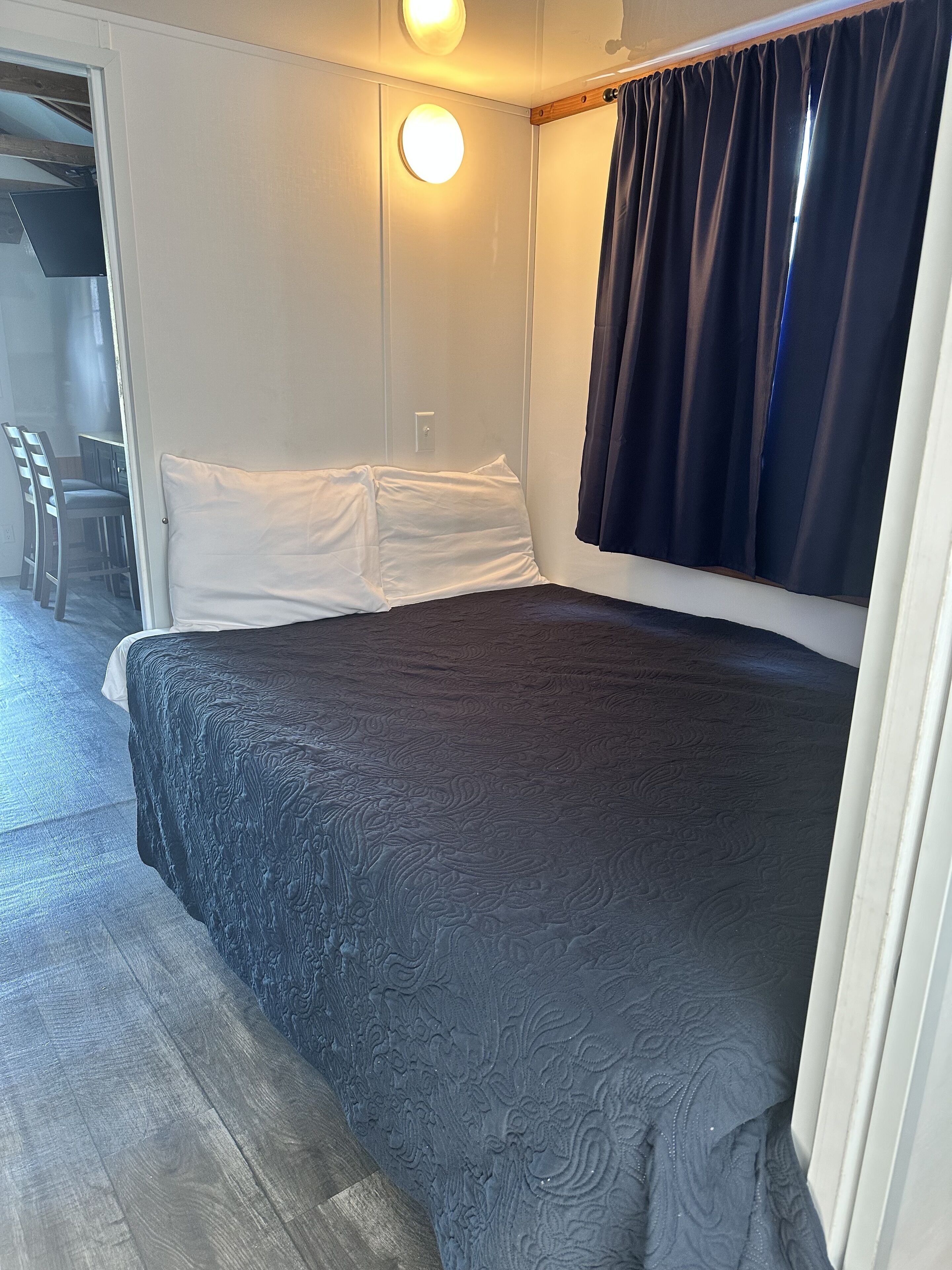A bedroom with a bed, white pillows, a wooden headboard, a round wall light, and a window with dark curtains.