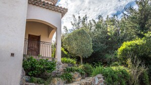 Un edificio con fachada de piedra, un balcón con barandilla de hierro forjado y un jardín exuberante con diversas plantas y un gran árbol.