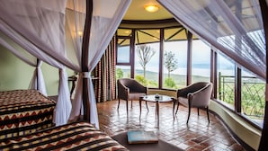 Quarto com cama, duas cadeiras, uma mesa pequena e uma janela grande com vista para a paisagem.