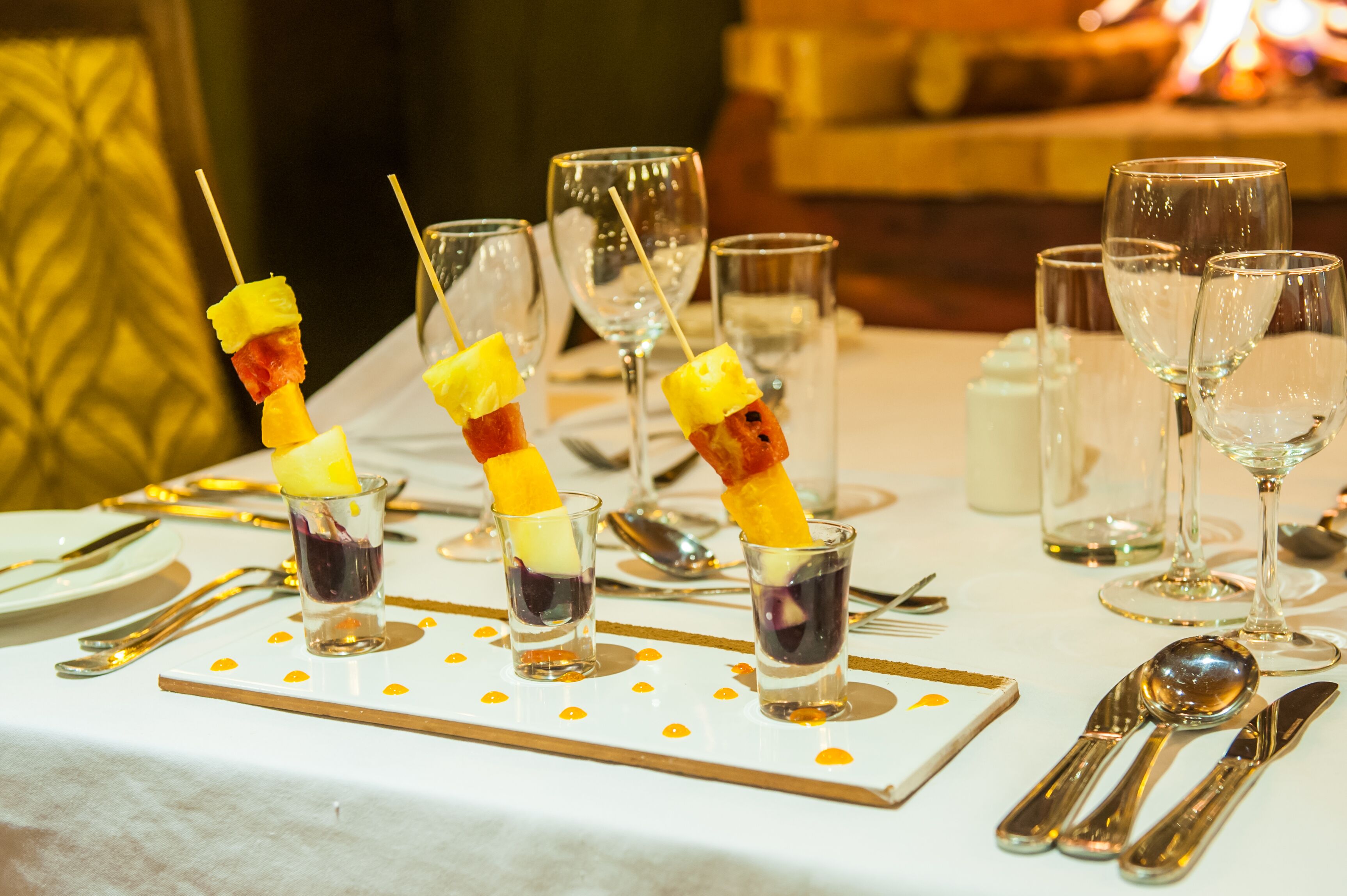 A table set with clear glasses, skewers with fruit, and a white tablecloth with gold accents.