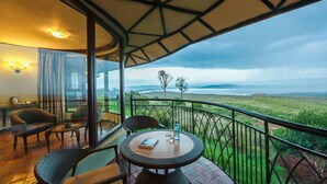 Uma varanda com mobiliário de vime, uma mesa com um livro e uma garrafa, e uma vista panorâmica de uma paisagem com árvores e colinas.