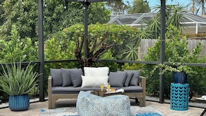 Un patio couvert aménagé avec un canapé, des coussins, un pouf à motifs et des plantes en pot.