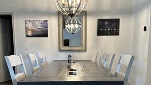 Une salle à manger avec une grande table en bois, des chaises blanches, un lustre et deux tableaux encadrés accrochés aux murs.