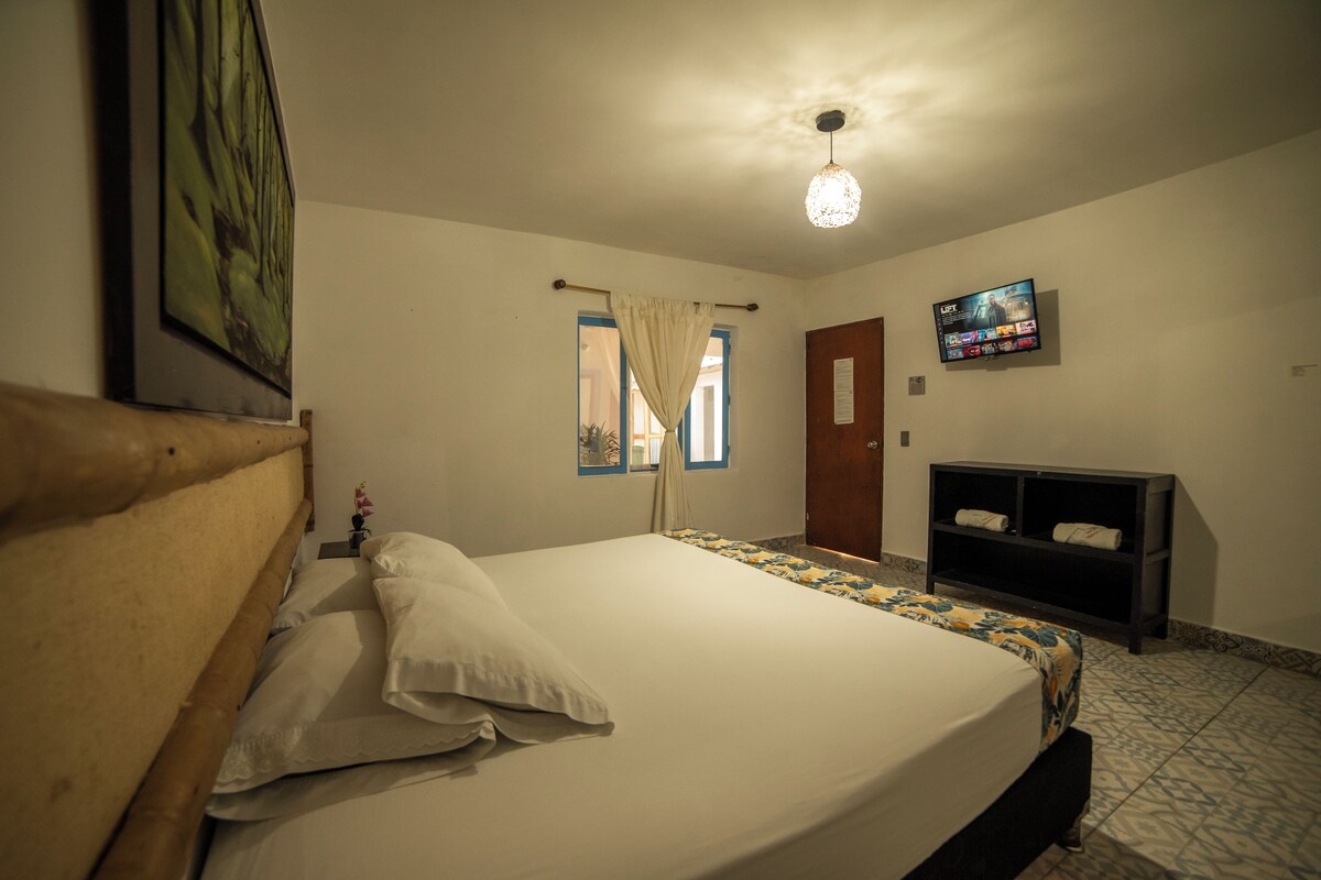 A bedroom with a bed, a TV, a wooden headboard, a window with curtains, and a shelf with towels.