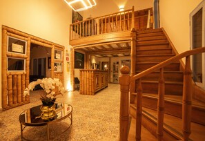 A wooden staircase leading to a second-floor balcony, a reception desk with a vase of white flowers, and a patterned floor.
