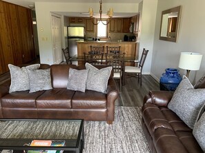 Een woonkamer met een bruine leren bank, een salontafel en een vloerkleed. In de achtergrond is een keuken met houten kasten en een eethoek met stoelen.