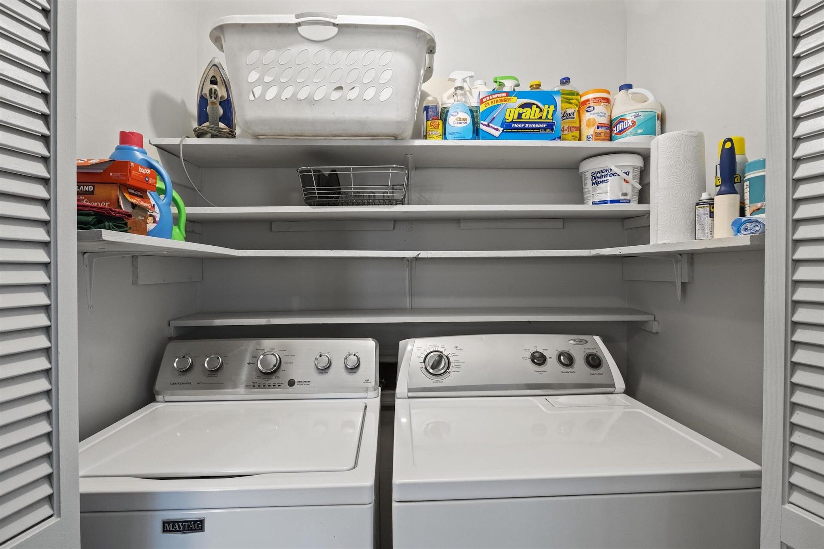 Pesuhuoneessa on kaksi Maytag-merkkiä olevaa pesu- ja kuivauskonetta, pyykkikori ja hyllyjä, joilla on säilytettynä siivoustarvikkeita.