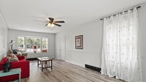 Een ruime kamer met een rode bank, een klein tafeltje, een plafondventilator en een raam met uitzicht op bomen.