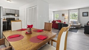 Een moderne keuken met witte kasten, een eethoek met een houten tafel en rode servetten, en een woonkamer met een rode bank en een lamp.