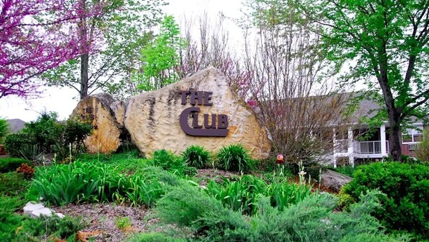 A large rock with 'THE CLUB' engraved on it, surrounded by lush greenery and trees.