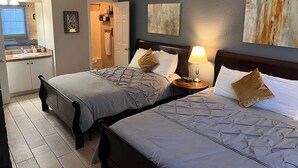 A hotel room with two beds, a bedside table with a lamp, a bathroom with a towel rack, and artwork on the walls.