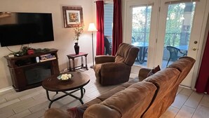 A living room with a sofa, armchair, and coffee table. There is a TV on the wall, a lamp, and a picture frame.