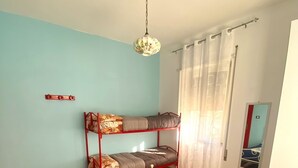 Una habitación con una cama individual, una litera, un colgador de luz, una ventana con cortinas y un pequeño espejo en la pared.