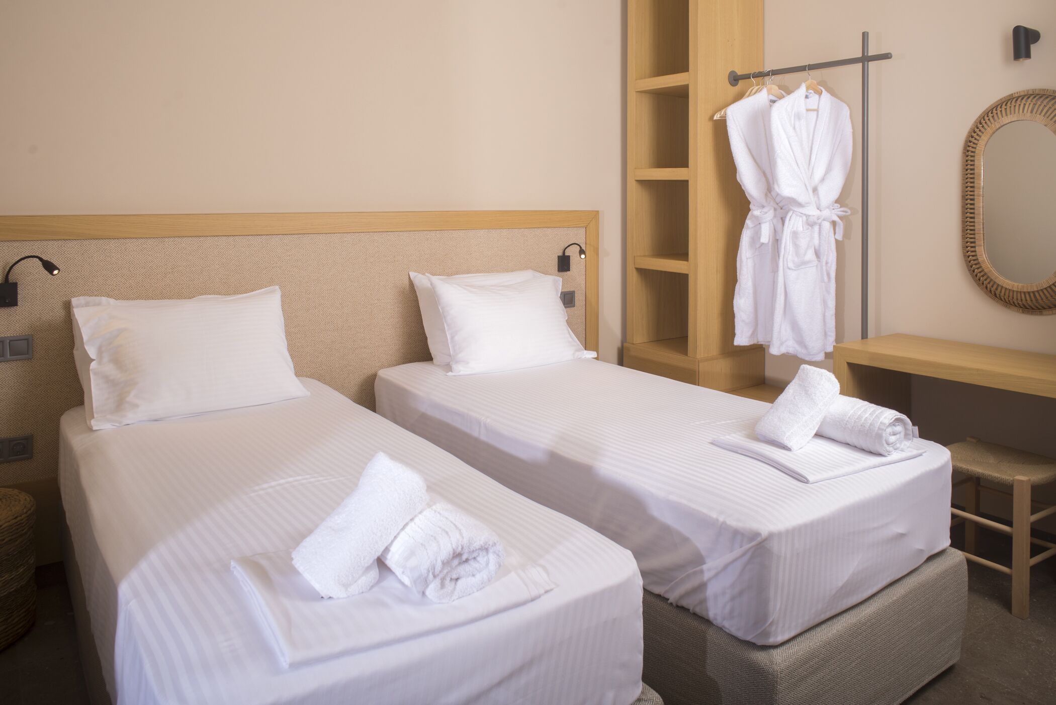 A hotel room with two beds, a wooden headboard, a bedside table, a robe rack, and a mirror.