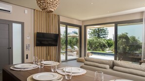 Un espace de repas moderne avec une table en bois pour quatre personnes, un téléviseur à écran plat fixé au mur et une grande porte-fenêtre coulissante donnant sur une terrasse extérieure avec une piscine et des chaises longues.