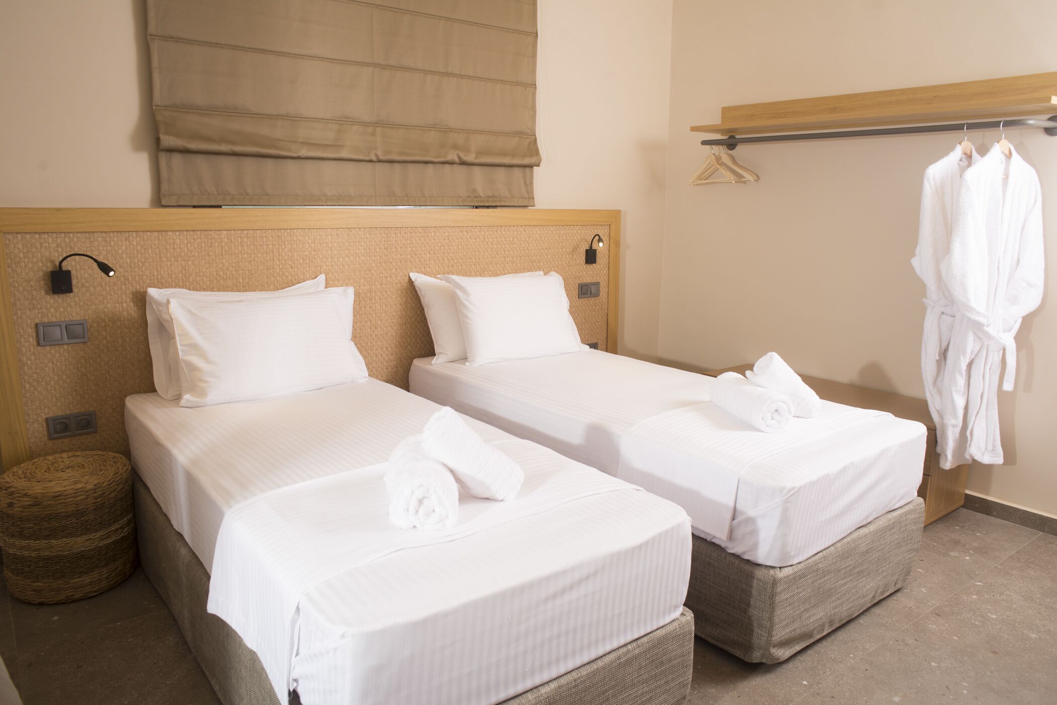 Habitación de hotel con dos camas individuales, una mesita de noche, un taburete redondo de mimbre, una bata colgada en un perchero y un ventanal grande con persiana color beige.