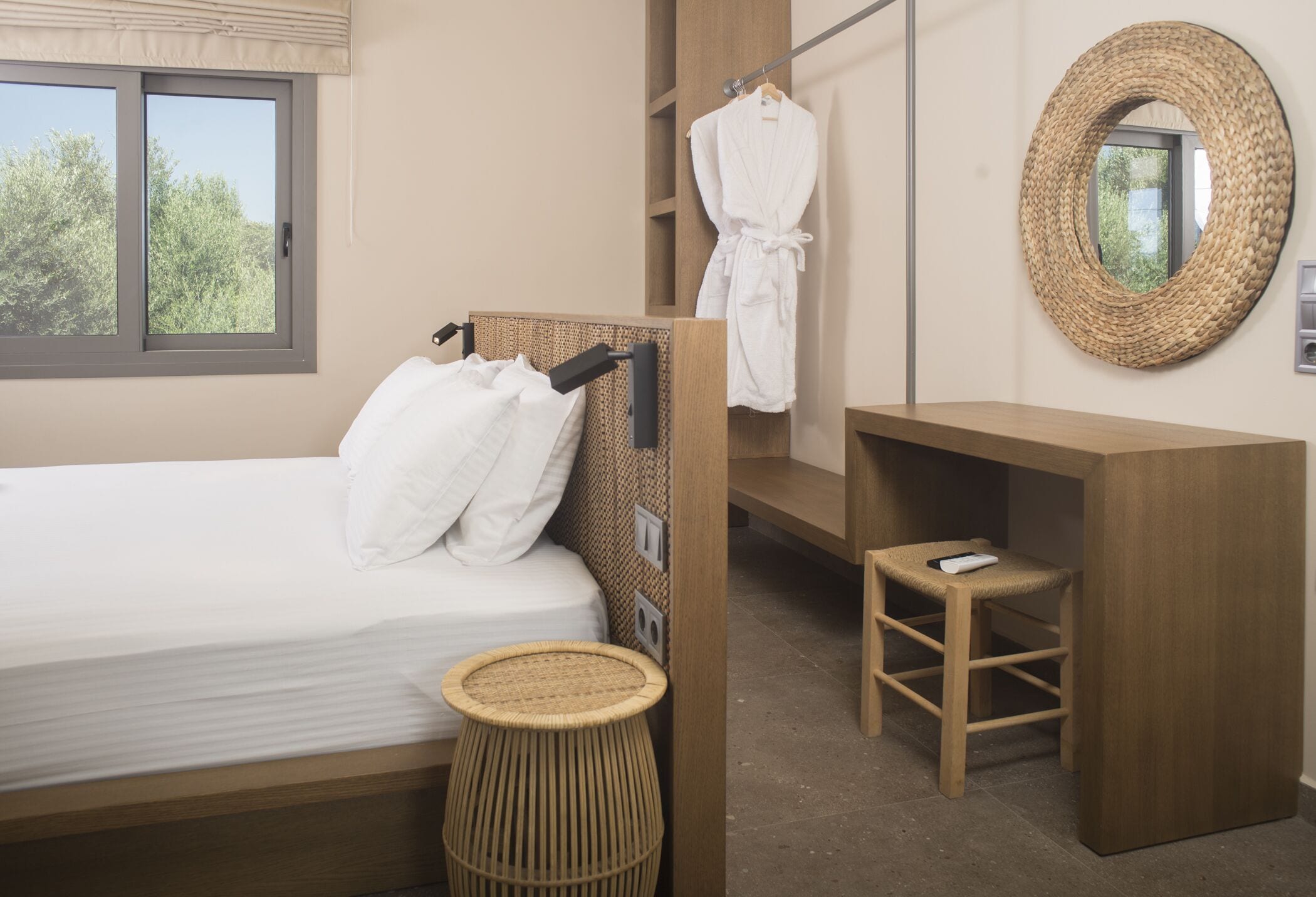 A hotel room with a bed, a wooden nightstand, a small stool, a hanging bathrobe, a woven mirror, and a window with a view of trees.