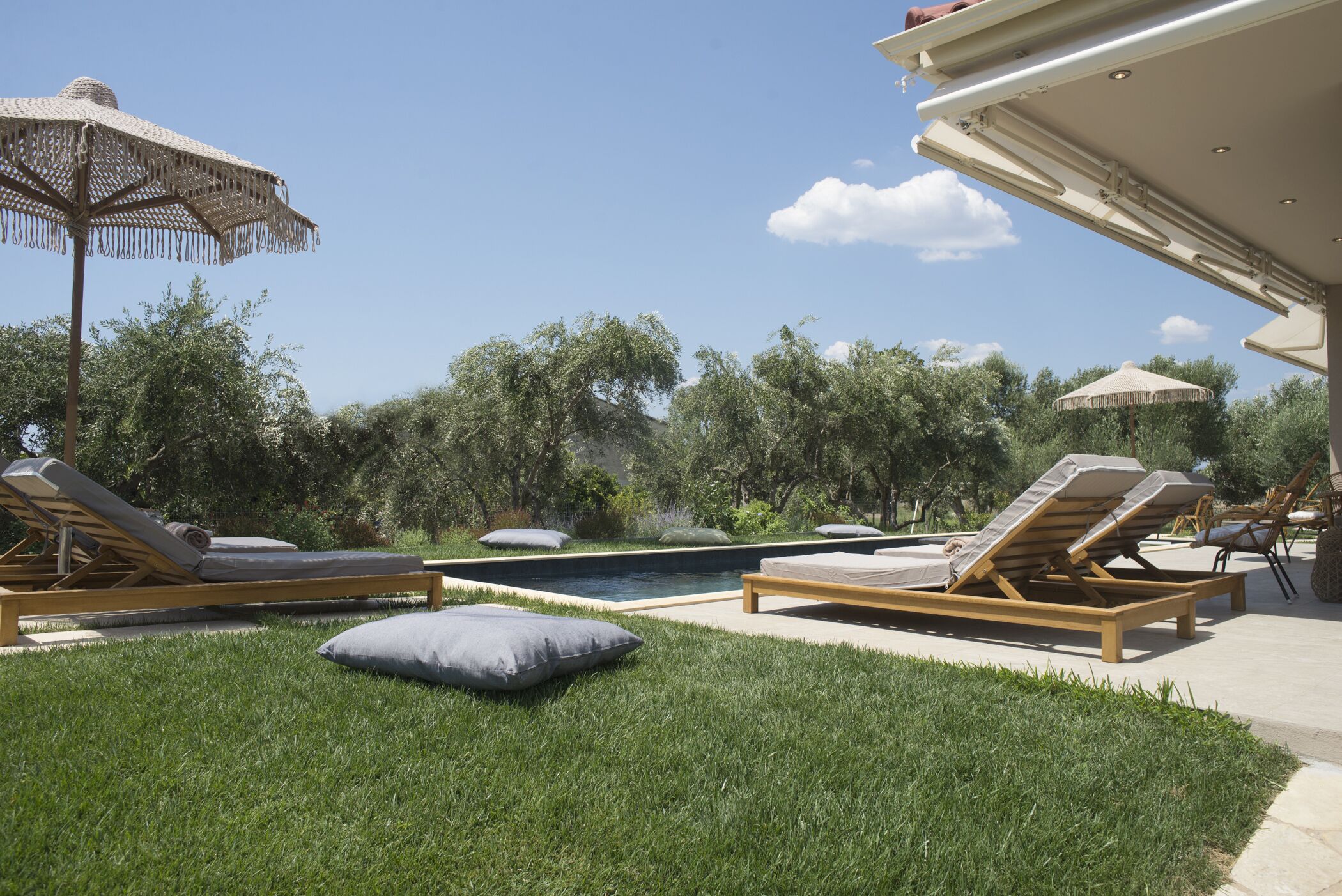 Zona de piscina amb gandules i ombrelles, envoltada de verdura i arbres.