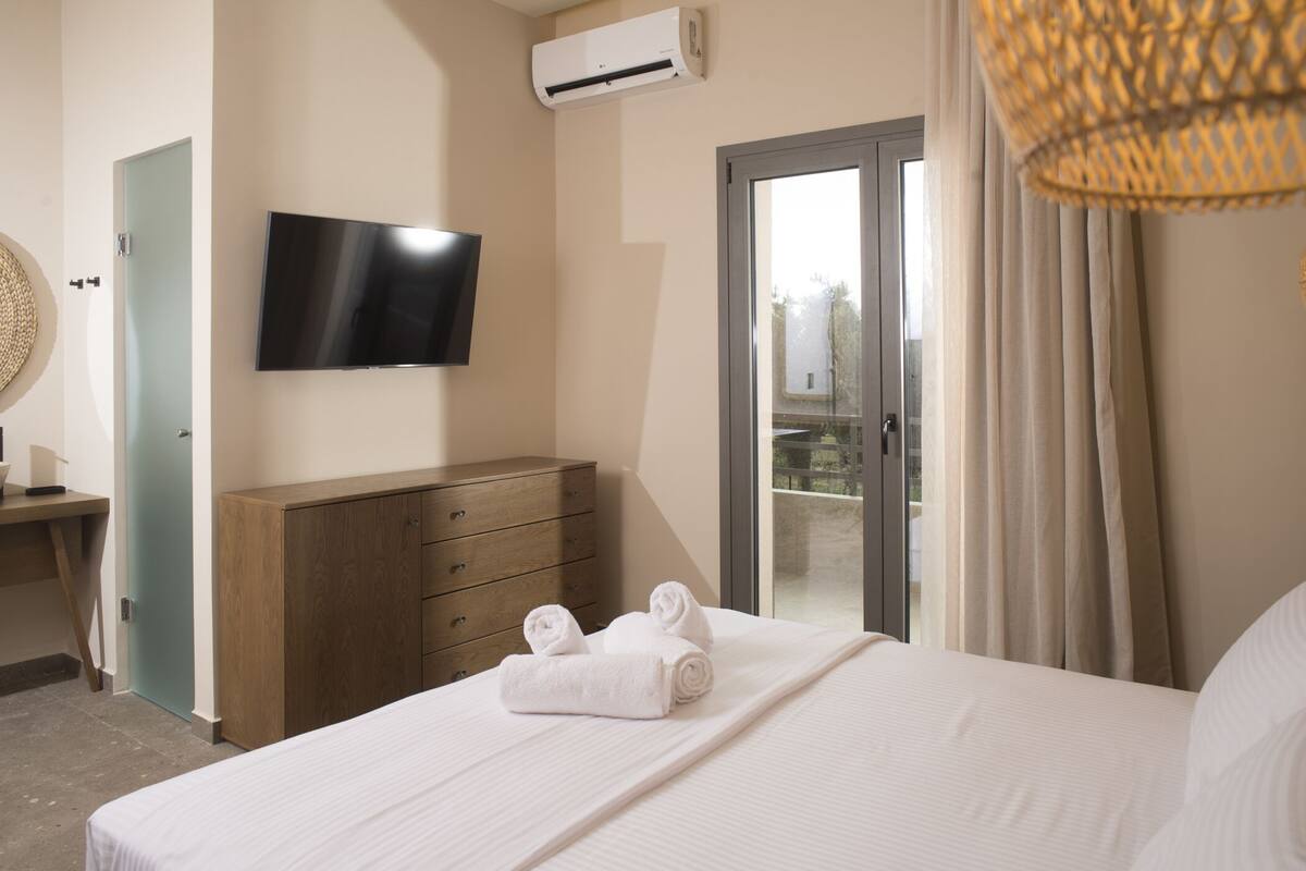 Quarto de hotel com cama, cómoda de madeira, TV de ecrã plano, unidade de ar condicionado e varanda com cortinas.