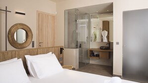 Une chambre d’hôtel avec un grand lit, une tête de lit en bois, un miroir rond, une douche en verre, une armoire en bois et un petit tabouret en osier.