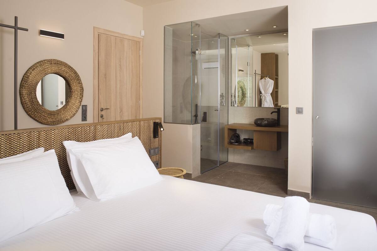 Quarto de hotel com cama grande, cabeceira de madeira, espelho redondo, chuveiro com divisória de vidro, guarda-roupa de madeira e um pequeno banquinho de vime.