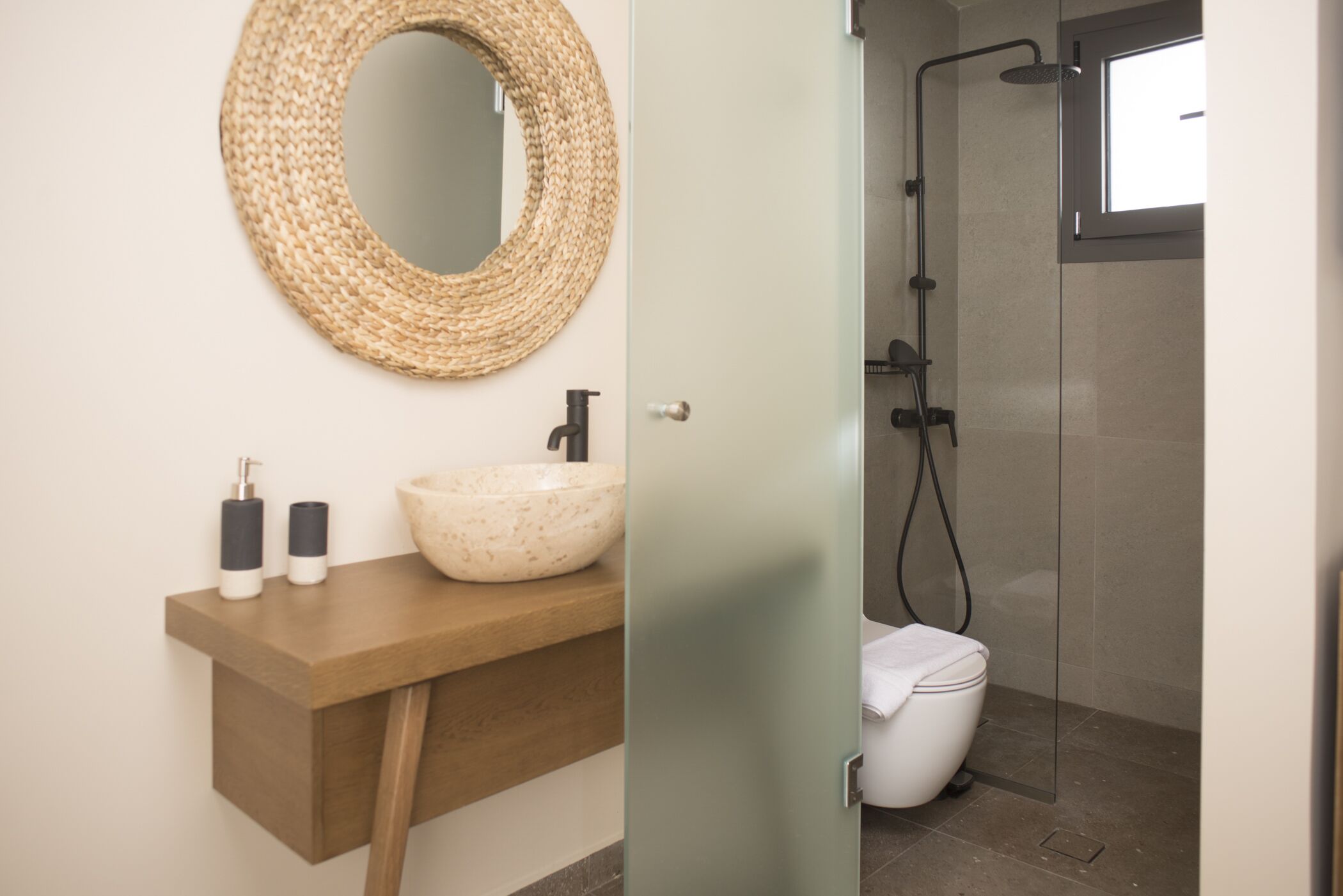 A bathroom with a round woven mirror, a wooden vanity, a bowl sink, and a glass-enclosed shower.